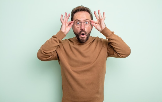 Middle age handsome man feeling shocked, amazed and surprised, holding glasses with astonished, disbelieving look