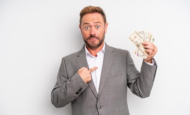 Middle age handsome man looking shocked and surprised with mouth wide open, pointing to self. dollar banknotes concept