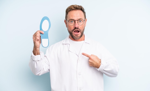 Middle age handsome man looking shocked and surprised with mouth wide open, pointing to self.  podiatrist concept