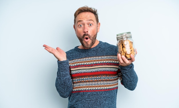 Middle age handsome man looking surprised and shocked, with jaw dropped holding an object. cookies bottle