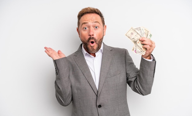 Middle age handsome man looking surprised and shocked, with jaw dropped holding an object. dollar banknotes concept