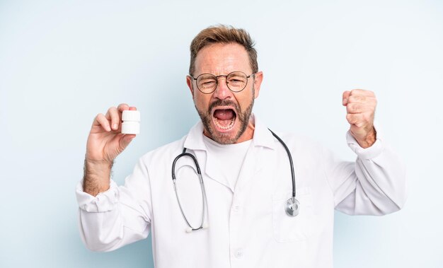 Middle age handsome man shouting aggressively with an angry expression. physician with pills bottle