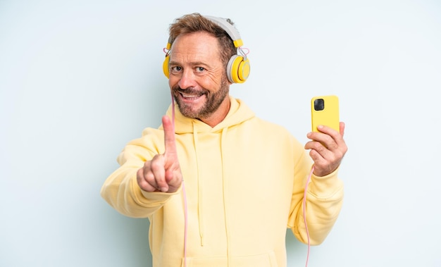 Middle age handsome man smiling proudly and confidently making number one. headphones and smartphone concept