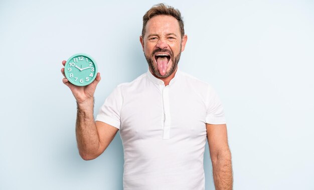 Middle age handsome man with cheerful and rebellious attitude, joking and sticking tongue out. alarm clock concept