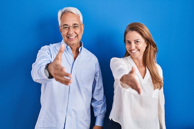 青い背景の上に立つ中年のヒスパニック系夫婦は、挨拶として握手を交わし、成功したビジネスを歓迎する笑顔を見せている