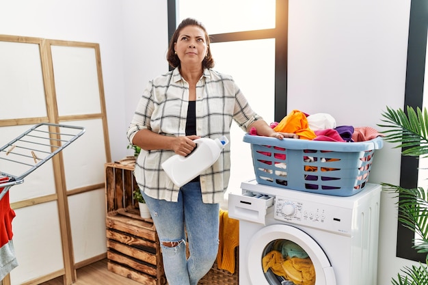 Middle age hispanic woman doing laundry holding detergent bottle smiling looking to the side and staring away thinking.