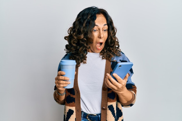 Middle age hispanic woman drinking a cup of coffee and looking at the smartphone screen celebrating crazy and amazed for success with open eyes screaming excited.