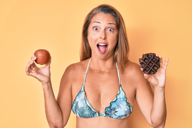 Middle age hispanic woman holding apple and donut celebrating crazy and amazed for success with open eyes screaming excited