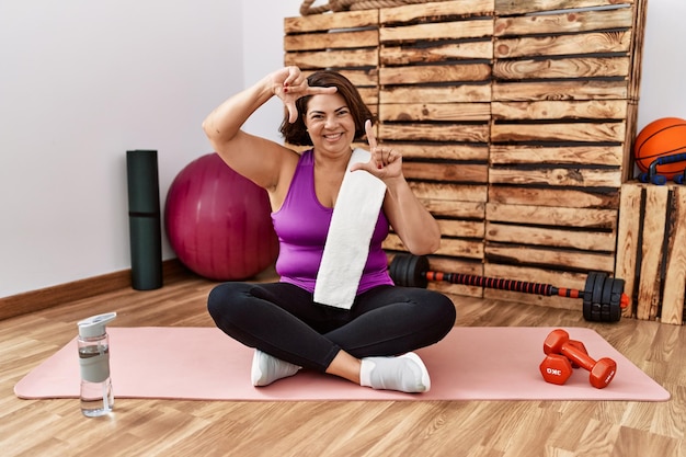 Middle age hispanic woman sitting on training mat at the gym smiling making frame with hands and fingers with happy face. creativity and photography concept.