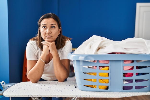 Middle age hispanic woman tired leaning on ironing board at laundry room