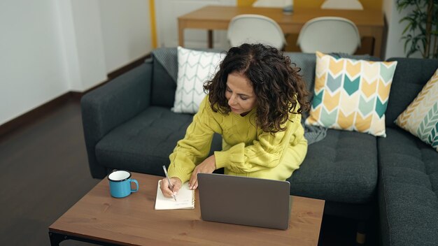 Middle age hispanic woman using laptop writing on notebook at home