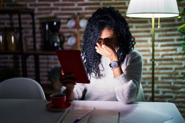 Foto donna ispanica di mezza età che utilizza il touchpad seduto sul tavolo di notte stanco sfregando il naso e gli occhi sentendosi affaticamento e mal di testa stress e concetto di frustrazione