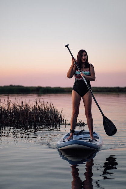 Middle age lady standing on blue sup board neat reeds holding\
oar in hands on lake looking away with pretty pink sky in\
background in evening active lifestyle for older people