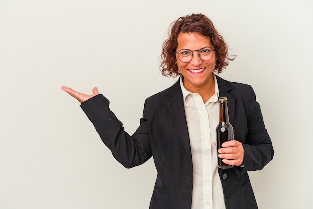 Middle age latin business woman holding a beer isolated on white background showing a copy space on a palm and holding another hand on waist.