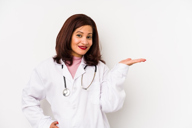 Middle age latin doctor woman isolated showing a copy space on a palm and holding another hand on waist.