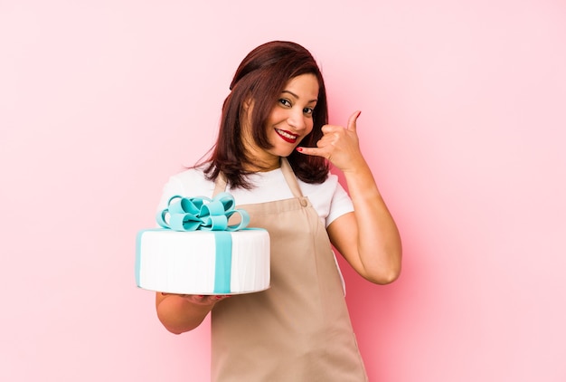 Middle age latin woman holding a cake isolated on a pink background showing a mobile phone call gesture with fingers.