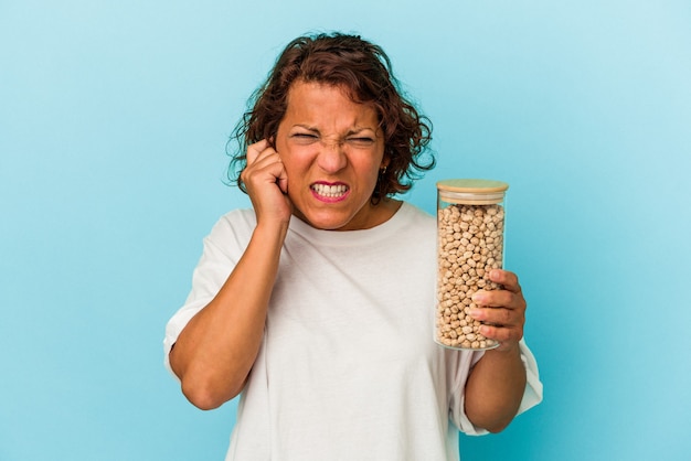 Middle age latin woman holding chickpeas jar isolated on blue background covering ears with hands.