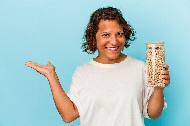 Middle age latin woman holding chickpeas jar isolated on blue background showing a copy space on a palm and holding another hand on waist.