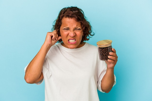 Middle age latin woman holding a coffee jar isolated on blue background covering ears with hands.