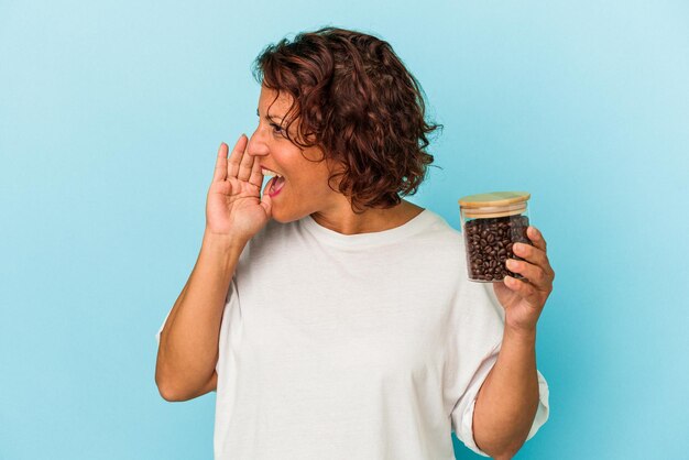 Middle age latin woman holding a coffee jar isolated on blue background shouting and holding palm near opened mouth.