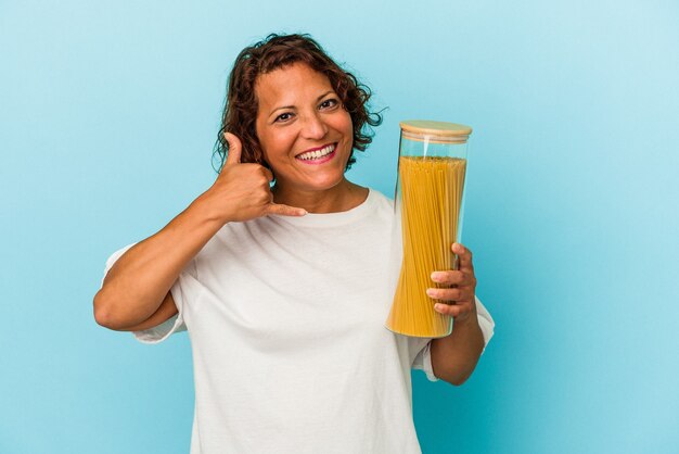 Middle age latin woman holding pasta jar isolated on blue background showing a mobile phone call gesture with fingers.