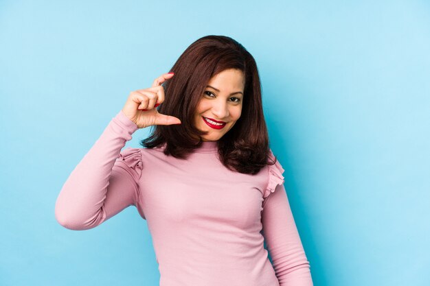 Middle age latin woman isolated holding something little with forefingers, smiling and confident.