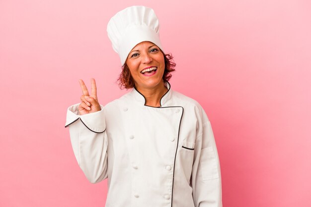 Middle age latin woman isolated on pink background joyful and carefree showing a peace symbol with fingers.