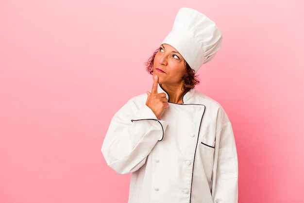 Donna latina di mezza età isolata su sfondo rosa guardando lateralmente con espressione dubbiosa e scettica.