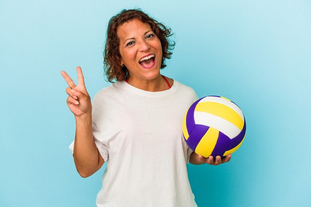 Middle age latin woman playing volleyball isolated on blue background joyful and carefree showing a peace symbol with fingers.