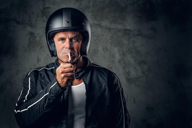 Middle age male in motorcycle helmet and leather jacket smoking a cigarette on grey background.