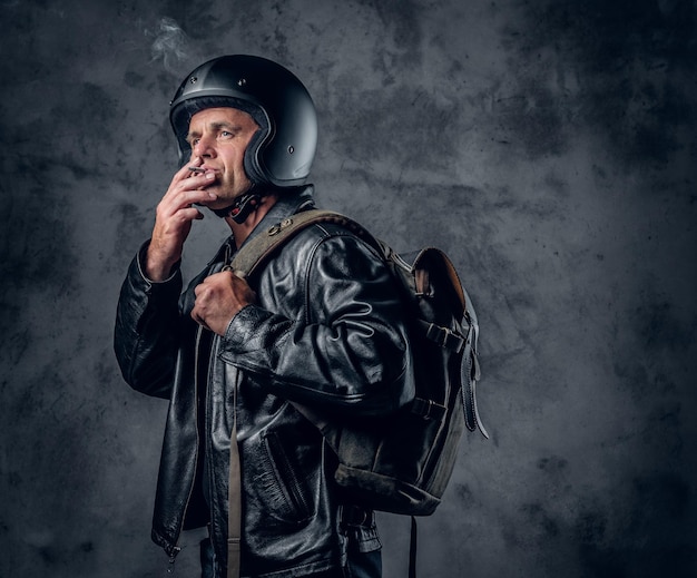 Middle age male in motorcycle helmet and leather jacket with rucksack smoking a cigarette on grey background.