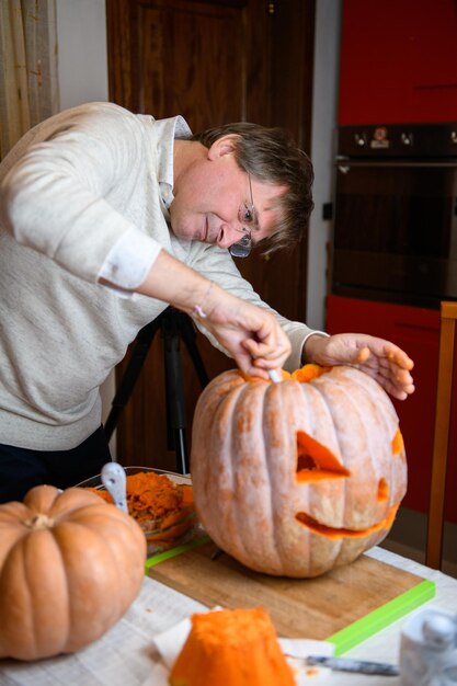 キッチンでハロウィーンの休日の装飾のためにジャッコランタンに大きなオレンジ色のカボチャを彫る中年男性の父