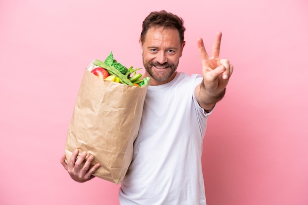 Middle age man holding a grocery shopping bag isolated on pink background smiling and showing victory sign