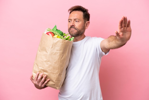 Middle age man holding a grocery shopping bag isolated on pink background