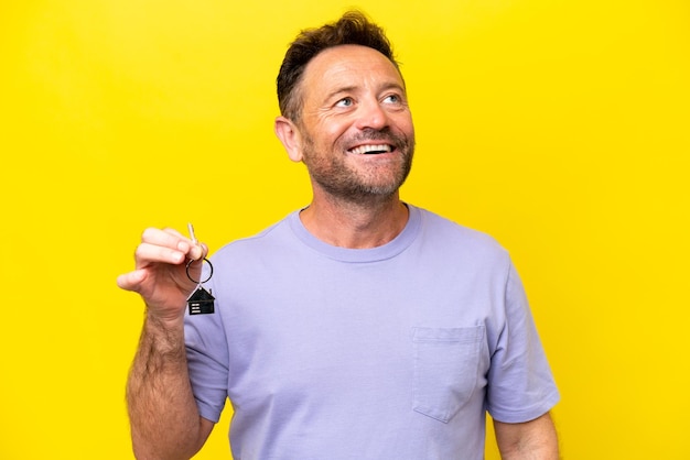 Middle age man holding home keys isolated on yellow background looking up while smiling