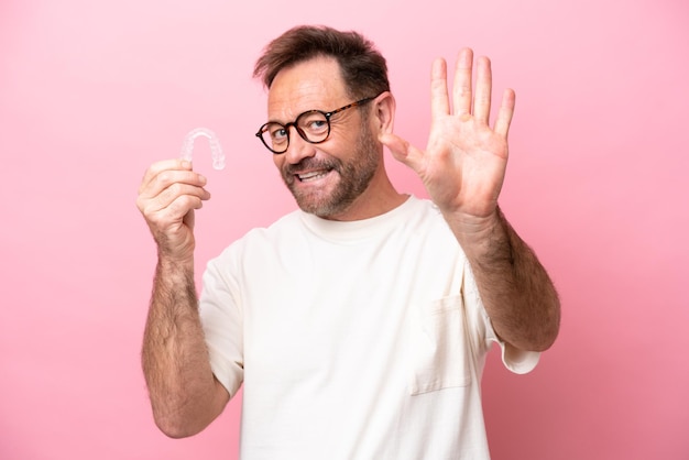 Middle age man holding invisible braces isolated on pink background saluting with hand with happy expression