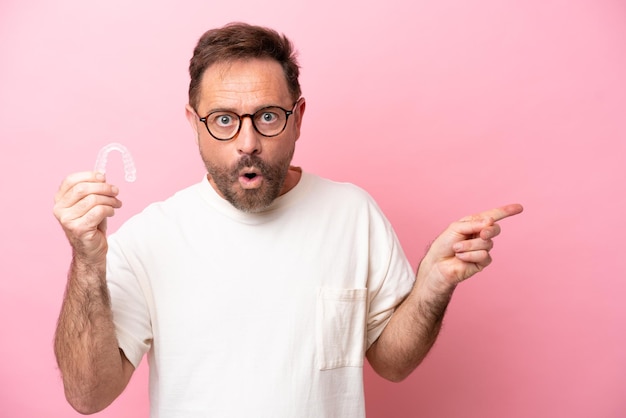 Middle age man holding invisible braces isolated on pink background surprised and pointing side