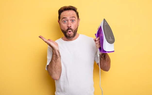 Middle age man looking surprised and shocked with jaw dropped holding an object clothes iron concept