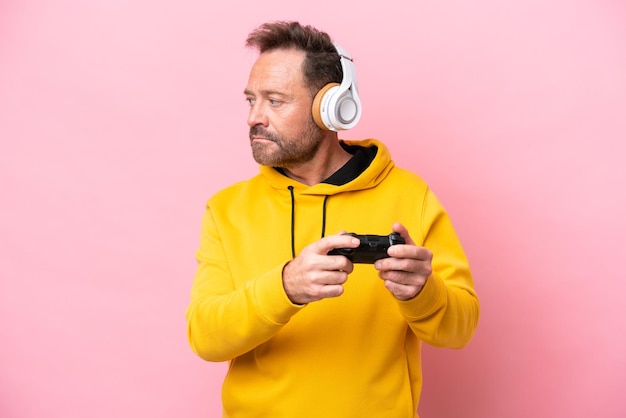 Middle age man playing with a video game controller isolated on pink background looking to the side