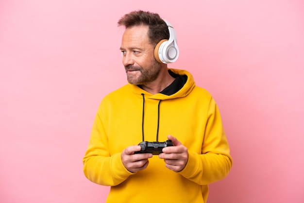 Middle age man playing with a video game controller isolated on pink background looking side