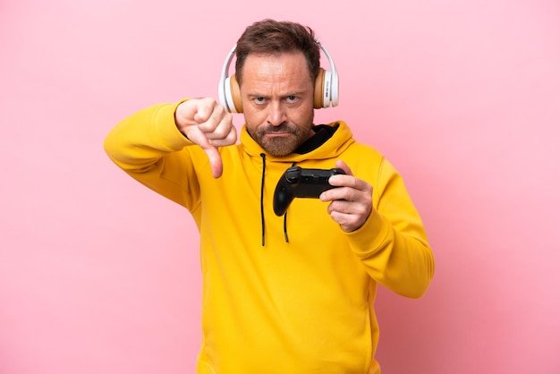 Middle age man playing with a video game controller isolated on pink background showing thumb down with negative expression