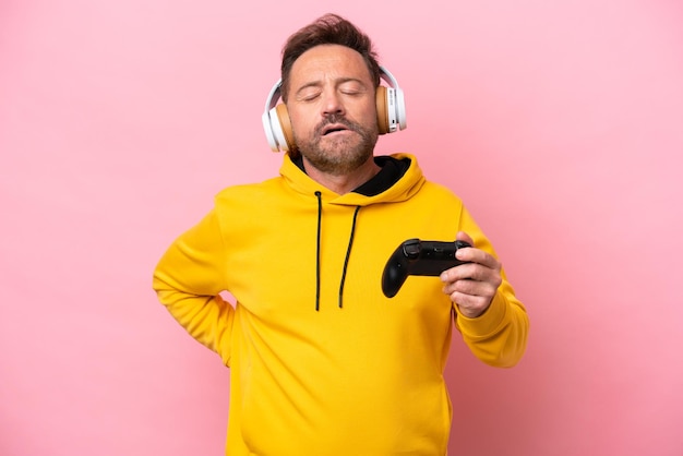 Middle age man playing with a video game controller isolated on pink background suffering from backache for having made an effort