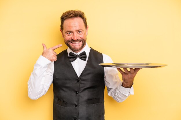 Middle age man smiling confidently pointing to own broad smile. waiter concept with tray