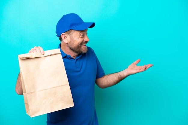 Мужчина средних лет берет сумку с едой на вынос, изолированную на синем фоне, с удивленным выражением лица, глядя в сторону
