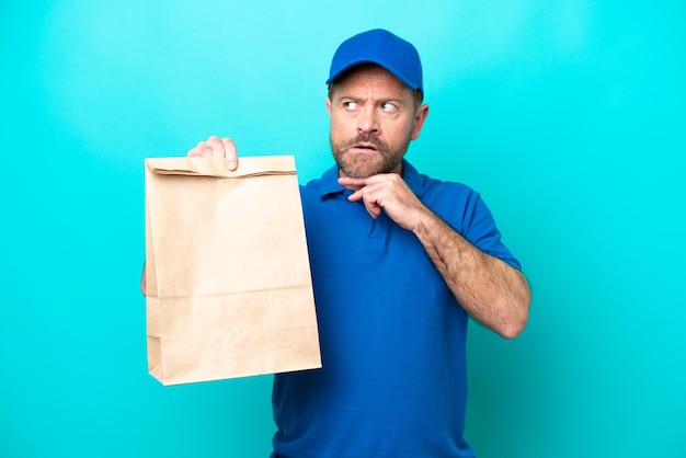 Мужчина средних лет берет сумку с едой на вынос, изолированную на синем фоне, сомневается и думает