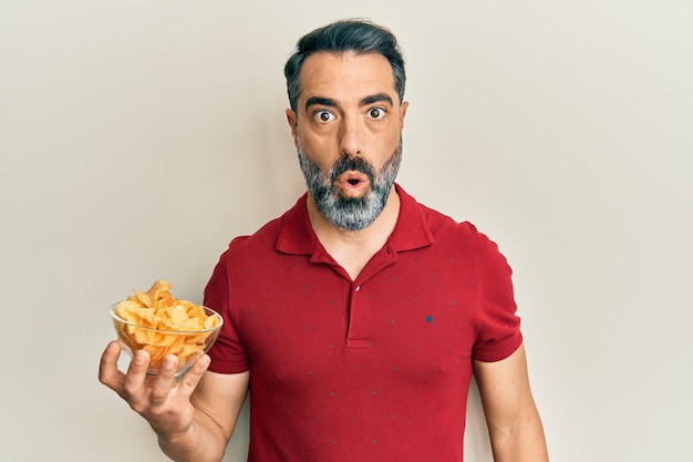 Middle age man with beard and grey hair holding potato chip scared and amazed with open mouth for surprise, disbelief face