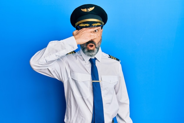 Middle age man with beard and grey hair wearing airplane pilot uniform peeking in shock covering face and eyes with hand, looking through fingers with embarrassed expression.