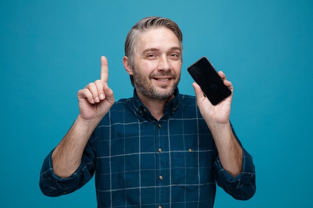 Foto uomo di mezza età con i capelli grigi in camicia di colore scuro che tiene lo smartphone puntato con il dito indice in alto felice e positivo guardando la fotocamera sorridente in piedi su sfondo blu