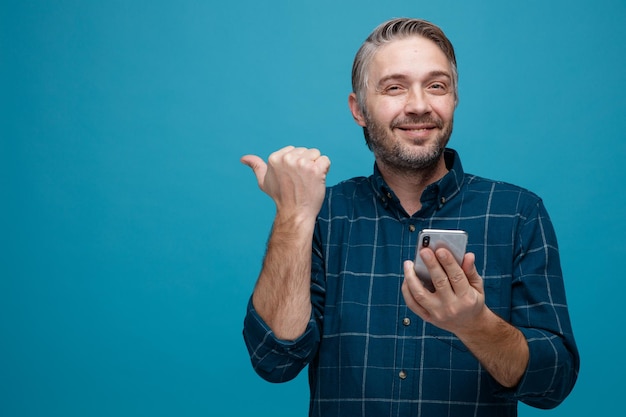 Uomo di mezza età con capelli grigi in camicia di colore scuro che tiene smartphone puntato con il pollice di lato guardando la fotocamera sorridente felice e positivo in piedi su sfondo blu