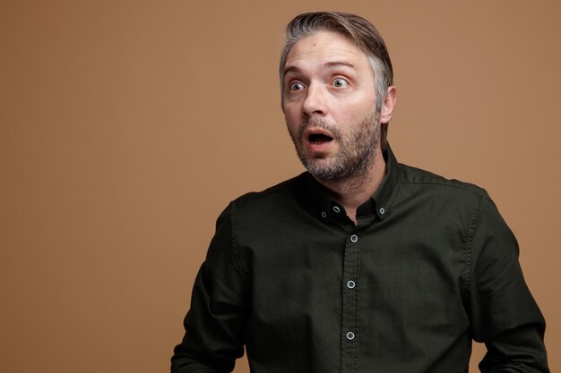 Middle age man with grey hair in dark color shirt looking aside amazed and shocked standing over brown background